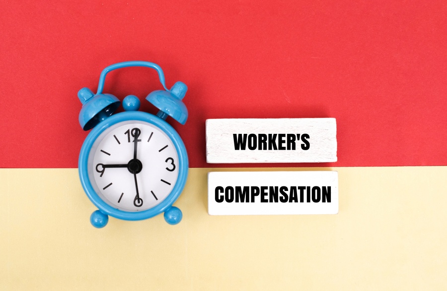 A blue alarm clock next to wooden blocks with text WORKER'S COMPENSATION on a colorful background. Concept for workplace injury insurance